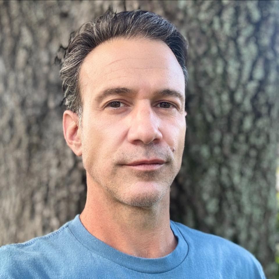 Justin Zoccolillo — a man with dark hair and facial hair wearing a blue shirt against a textured stone wall.